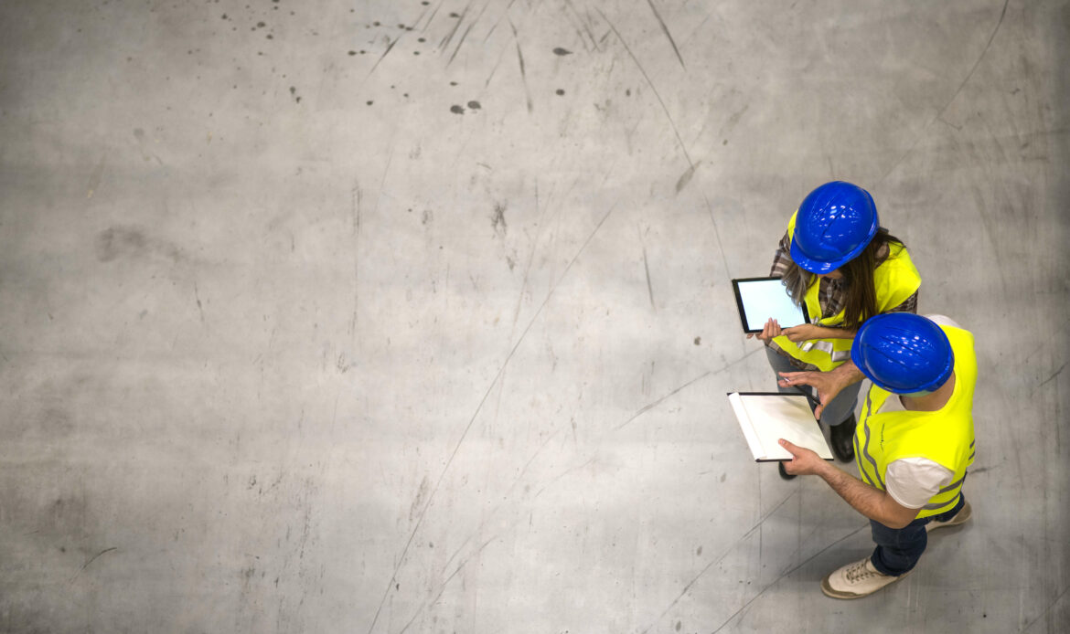 Top view of two industrial workers wearing hardhats and reflective jackets holding tablet and checklist on gray concrete background.
